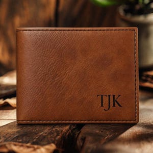 May include: A brown leather wallet with the initials "TJK" embossed on the lower right corner. The wallet has a textured surface and is displayed on a wooden surface.