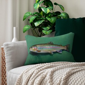 May include: A rectangular decorative pillow featuring a detailed illustration of a rainbow trout. The fish has iridescent scales and is set against a solid green background. The pillow is on a gray sofa with a cream-colored throw.
