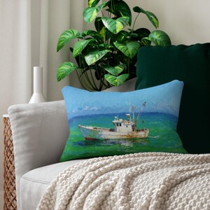 May include: A rectangular throw pillow featuring a painting of a white fishing boat on turquoise water under a blue sky. The pillow is on a gray sofa with a cream-colored blanket. A green plant and dark green pillow are in the background.