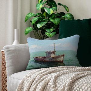 May include: A rectangular decorative pillow featuring a painting of a weathered fishing boat on the sea. The boat is gray and brown, set against a blue sky and turquoise water. The pillow is on a gray sofa with a cream-colored throw.