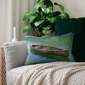 May include: Decorative pillow featuring a serene image of a wooden canoe floating on calm, turquoise water. The pillow has a rectangular shape and is placed on a light gray sofa, with a green plant and pillows in the background.