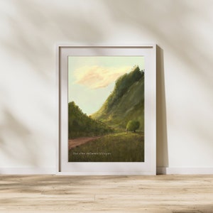 May include: A framed painting of a green mountain with a dirt road leading up to it. The sky is a light blue with a few white clouds. The text "the view between villages" is at the bottom of the painting.