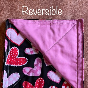 May include: Reversible quilted square with a black background and a pink background. One side features a pattern of red, pink, and white hearts. The other side is solid pink. The text "Reversible" is printed above the square.
