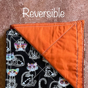 May include: Reversible quilt with a black background and a pattern of colorful sugar skull cats. The other side of the quilt is solid orange.