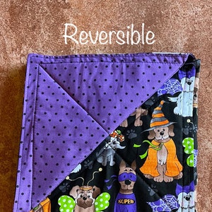 May include: Reversible quilted square with a purple polka dot fabric on one side and a black fabric with a Halloween dog theme on the other side. The dog designs include a dog wearing a witch hat, a dog wearing a superhero costume, a dog with angel wings, and a dog with a spider web.