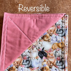 May include: Reversible pink and white fabric square with a pattern of bunnies wearing flower crowns. The fabric is sewn together with a diagonal seam.