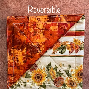 May include: Reversible placemat with two designs. One side features a brown and orange abstract pattern. The other side features a white background with a floral pattern of sunflowers, daisies, and leaves.