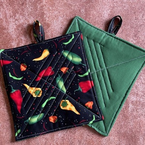 May include: Two square potholders. One is black with a pattern of red, yellow, and green chili peppers. The other is solid green. Both have a loop for hanging and white stitching.