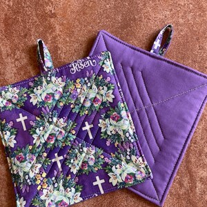 May include: Two purple pot holders. One is plain and the other has a floral pattern with white crosses and the word "Risen" in white lettering.