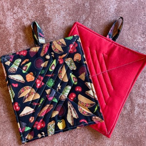 May include: Two pot holders, one red and one black with a pattern of tacos, peppers, and other Mexican food.