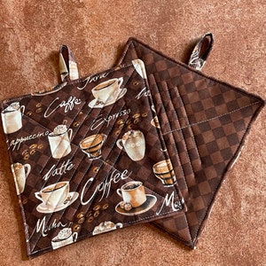 May include: Two brown quilted pot holders with a checkered pattern on one side and a coffee themed print on the other. The print features various types of coffee drinks, including cappuccino, latte, mocha, espresso, and java, with illustrations of coffee cups and beans.