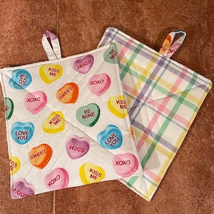 May include: Two white cotton pot holders with a quilted design. One pot holder has a pink, yellow, and blue plaid pattern. The other pot holder has a white background with a repeating pattern of pink, red, orange, yellow, green, and blue hearts with various text on them, including "Love You", "Kiss Me", "Hugs", "Sweet", "Be Mine", and "XOXO".