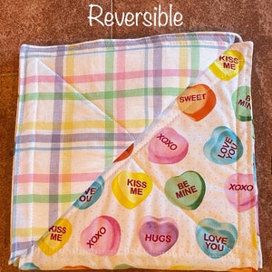 May include: Reversible quilted hot pad with a plaid pattern on one side and a white background with colorful heart-shaped designs on the other side. The hearts have various sayings on them, including "Kiss Me", "Love You", "Be Mine", "Hugs", "Sweet", and "XOXO".