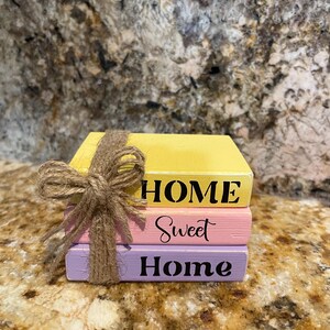 May include: A stack of three wooden blocks painted yellow, pink, and purple, tied with twine. The top block reads "HOME", the middle block reads "Sweet", and the bottom block reads "Home" in black lettering. The blocks are on a speckled surface.