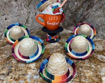 May include: A set of six miniature straw sombreros with colorful trim, arranged around a small orange mug with the text "Let's fiesta!" on it. The mug is topped with a stack of three sombreros and a straw.