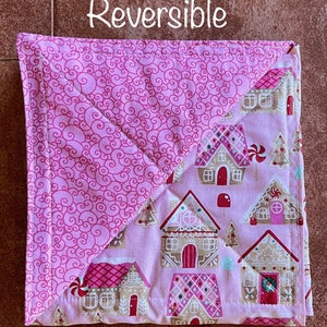 May include: Reversible pink and white fabric square with a gingerbread house pattern on one side and a pink swirl pattern on the other side. The text "Reversible" is written in white on a brown background above the square.