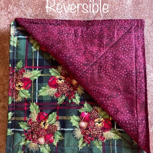 May include: Reversible quilted placemat with a green plaid pattern on one side and a burgundy dotted pattern on the other side. The plaid side features a festive design of holly, pine cones, and bells.