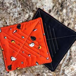 May include: Two quilted pot holders, one orange with a white ghost and star pattern and one black with white stitching.