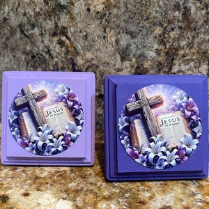 May include: Two square, painted wooden blocks, one light purple and one dark purple. Each block features a circular image with a wooden cross, an open book, and the text "JESUS HE IS RISEN" surrounded by purple and white flowers.