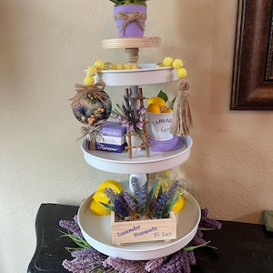May include: A three-tiered decorative tray with a lavender and lemon theme. The top tier features a purple pot with lemons and white flowers. The middle tier has lavender and a "Lemon 5 Cent" cup. The bottom tier has a wooden box of lavender bouquets.
