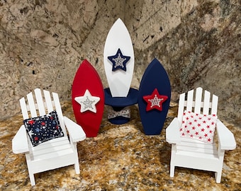 Pode incluir: Um conjunto decorativo com tema patriótico com duas cadeiras Adirondack brancas com almofadas com estampa de estrelas, três ornamentos em forma de prancha de surf em vermelho, branco e azul e enfeites de estrelas. O conjunto evoca um tema de verão ou praia.