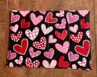 May include: Black fabric placemat with a repeating pattern of pink and red hearts. The hearts have different patterns, including polka dots, stripes, and gingham.