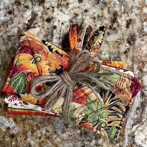 May include: Set of two fall-themed oven mitts with a pumpkin, leaf, and wheat design. The mitts are tied together with twine.