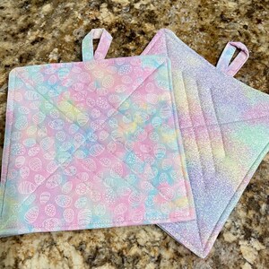 May include: Two pastel-colored quilted pot holders. One is pink and blue with a white egg pattern. The other is a light purple and yellow ombre.