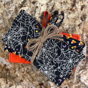 May include: Set of four black and orange Halloween-themed pot holders with a spiderweb design. The pot holders are tied together with twine.