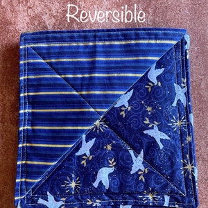 May include: Reversible square fabric item with a blue and gold striped pattern on one side and a blue dove and star pattern on the other. The text "Reversible" is at the top. The item is folded and stitched with white thread.
