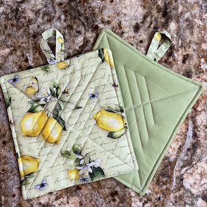 May include: Two green quilted pot holders. One is plain and the other has a lemon and floral print with handwritten text. Both have a loop handle.