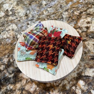 May include: A collection of small, square fabric sachets with various autumn-themed patterns, including plaid, houndstooth, and leaf prints. The sachets are arranged on a round, light-colored wooden base.