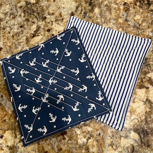May include: Two square quilted pot holders. One is navy blue with white anchors and stars. The other has white and navy blue vertical stripes. Both have white stitching.