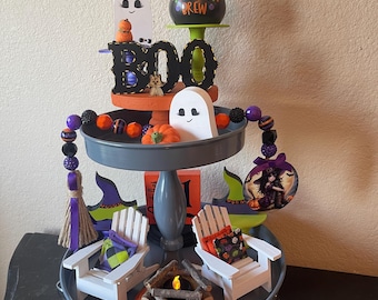 May include: A three-tiered Halloween-themed tray with decorative elements. The top tier features a witch's hat, a cauldron with "Witches Brew" text, and a "BOO" sign. The middle tier has ghost figures and pumpkins. The bottom tier includes miniature chairs, a campfire, and a broom.