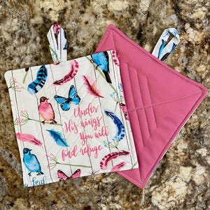 May include: A pair of quilted pot holders. One side is pink with a quilted design. The other side is white with a pattern of feathers, birds, butterflies, and the text "Under His wings you will find refuge".