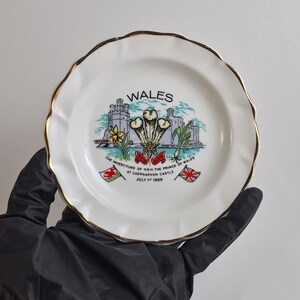 May include: A white ceramic commemorative plate with a scalloped edge and gold trim. The center features a graphic of Caernarvon Castle, the Prince of Wales feathers, and text reading WALES THE INVESTITURE OF H.R.H. THE PRINCE OF WALES AT CAERNARVON CASTLE JULY 1ST 1969.