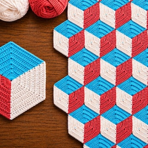 Peut inclure: Gros plan sur un motif de crochet fait à la main en rouge, blanc et bleu. Le motif crée un effet de cube 3D. L'image montre également des pelotes de laine bleues, blanches et rouges, et un seul cube crocheté.