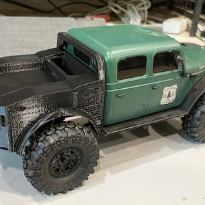 Puede incluir: Un modelo de camión verde y negro con neumáticos grandes y texturizados. El camión tiene una cabina verde con una caja y parte delantera negras. Un emblema blanco con un árbol está en la puerta. El modelo está sobre una superficie blanca.