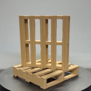 Könnte beinhalten: Zwei kleine Holzpaletten, eine auf der anderen gestapelt. Die Paletten sind hellbraun und bestehen aus vertikalen und horizontalen Holzlatten. Auf der unteren Palette sind die Buchstaben "JD3" aufgedruckt. Der Hintergrund ist neutralgrau.