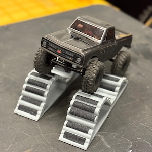 Puede incluir: Una camioneta Chevrolet negra en miniatura con neumáticos grandes y con tacos está colocada sobre una estructura de rampa blanca y gris. La camioneta tiene una parrilla delantera detallada y un pequeño emblema rojo. La rampa tiene una serie de escalones con superficies texturizadas negras.