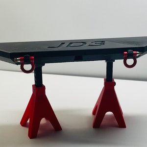 Może przedstawiać: Czarny, kanciasty przedmiot z napisem "JD3" jest podparty przez dwa czerwone stojaki. Obiekt posiada dwa czerwone pierścienie typu D. Stojaki są trójkątne i mają po trzy nogi. Tło jest białe.