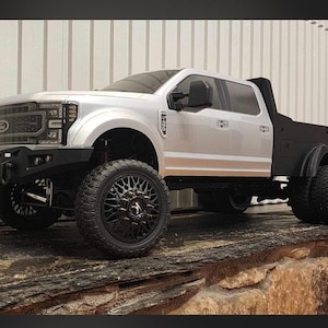 May include: A silver and black model pickup truck with large black wheels and a black flatbed. The truck has a detailed front grille, a black front bumper, and a black side mirror. The truck is sitting on a wooden surface.