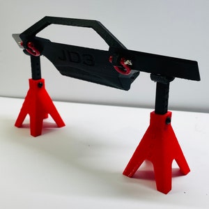 May include: A black 3D-printed model of a car door, supported by two red jack stands. The door has the text "JD3" printed on it and is connected to the stands with red loops.