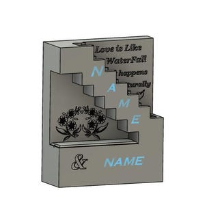 Op de afbeelding: Een grijs, driedimensionaal decoratief blok met een trapontwerp. Het blok bevat het woord "NAME" in blauw, samen met de zin "Love is Like Waterfall happens naturally." Bloemenontwerpen en een ampersand zijn ook aanwezig.