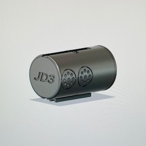 以下が含まれることがあります： 片方の端に「JD3」の文字が入った、円筒形の濃いグレーの物体。この物体には、小さな穴のある円形のデザインが2組あり、上部にスロットがあります。長方形のベースの上にあります。