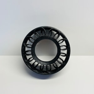May include: A black, circular decorative object with an open center. The outer edge features a repeating pattern of interconnected, oval-shaped designs. The object is made of a matte material and sits against a white background.