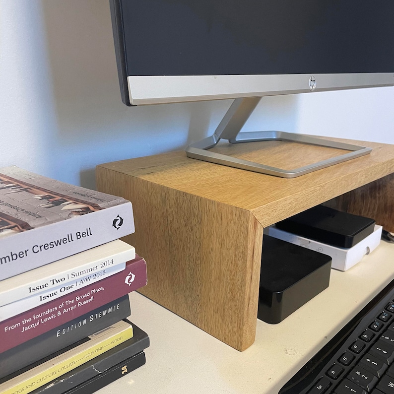 May include: A wooden monitor stand with a curved front edge. The stand is holding a computer monitor and has two black boxes underneath. A stack of books with colorful covers is on the left side of the stand.