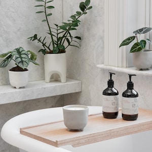 May include: A white bathtub with a wooden tray on top. The tray has a grey concrete candle with three legs and two bottles of body wash. The bottles are labelled "north & grove" and have a floral design. The bottles are on a white shelf with two potted plants.