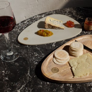 May include: Two serving trays, one white and one wooden, with various snacks. The white tray has a blue cheese wedge, a small bowl of yellow relish, and a dollop of red jam. The wooden tray has a stack of round crackers, a slice of cheese, and a gold coin-shaped decorative element.