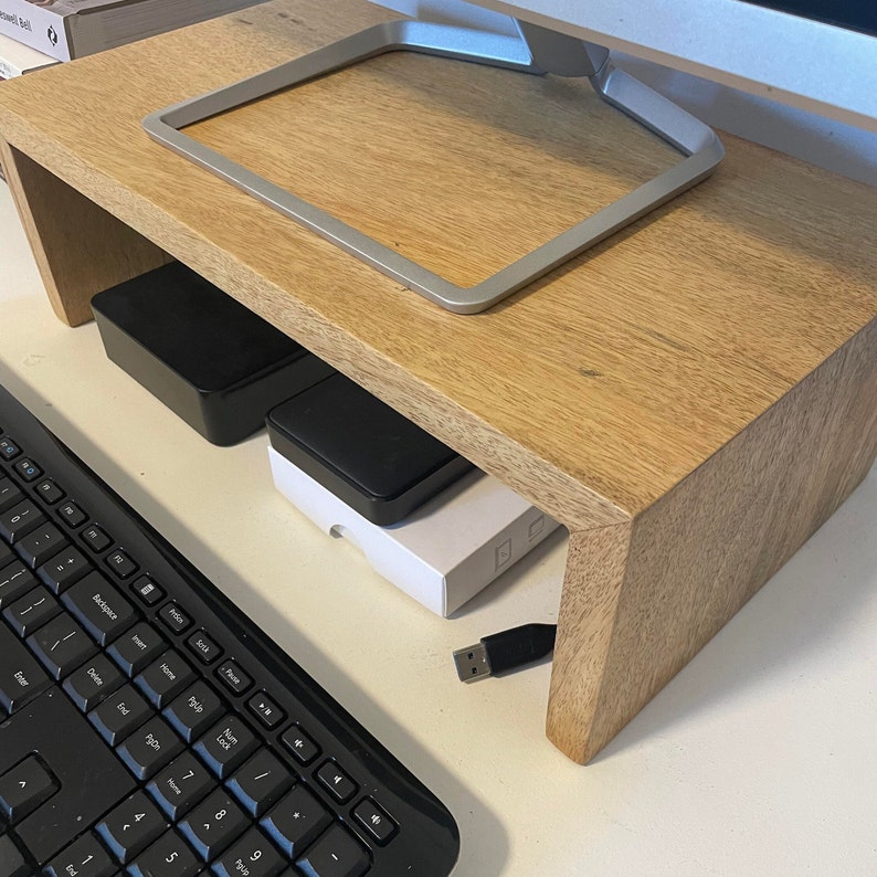 May include: A wooden monitor stand with a light brown finish. The stand has a raised edge to hold a monitor and a lower shelf for storing items. A black keyboard and a black USB cable are visible in the image.